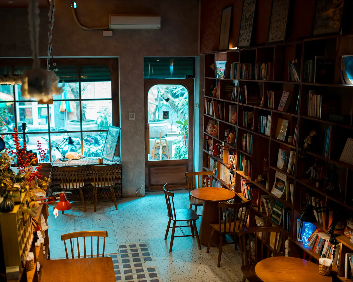 Cozy cafe interior with wooden tables and chairs, lined with bookshelves. Soft lighting creates a warm, inviting atmosphere. Large windows show lush plants outside.
