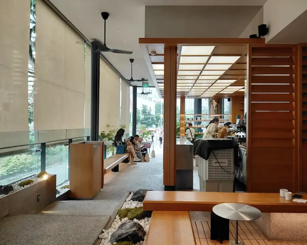 Modern cafe interior with wooden elements, soft lighting, and large windows. People sit along the wall, creating a relaxed and cozy atmosphere.