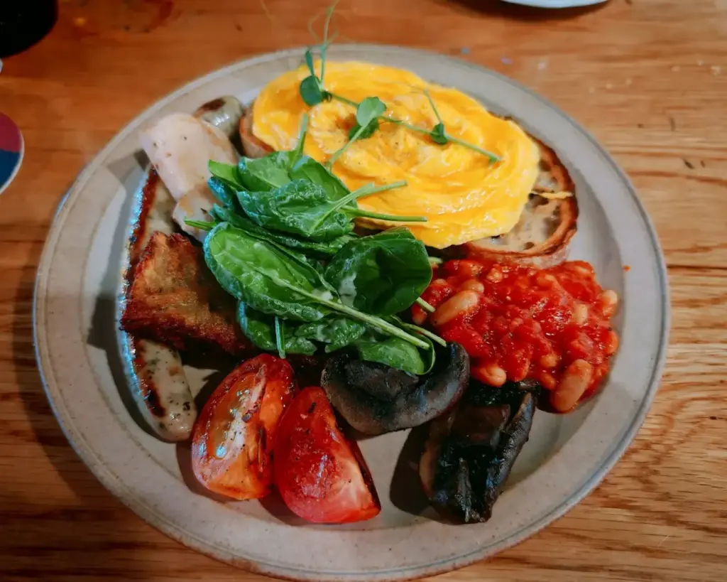 A hearty breakfast plate with scrambled eggs on toast, sausages, roasted tomato, spinach, mushrooms, baked beans, and toast, on a wooden table.