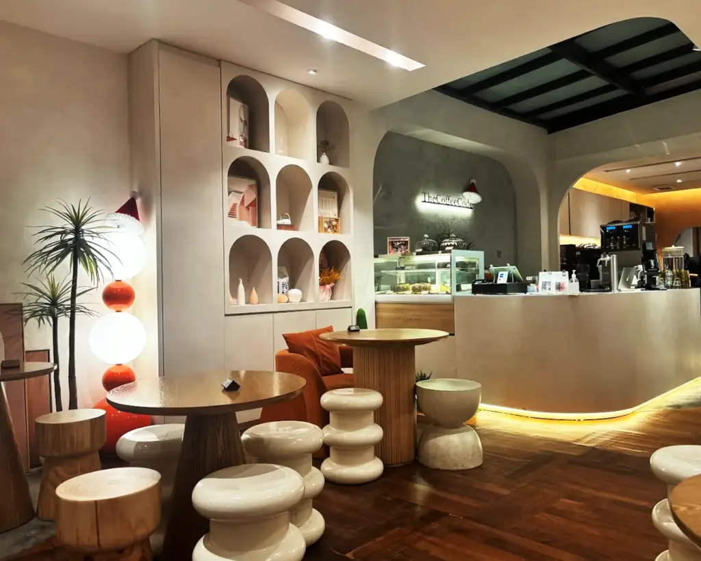 Cozy café interior with warm lighting, modern round tables, and sculptural chairs. Decorative shelves and a display case enhance the inviting atmosphere.