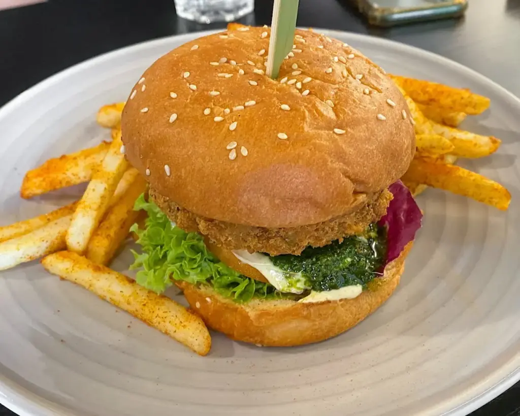 A sesame seed bun burger with crispy patty, lettuce, and sauce, served with seasoned fries on a plate. The scene suggests a casual dining experience.