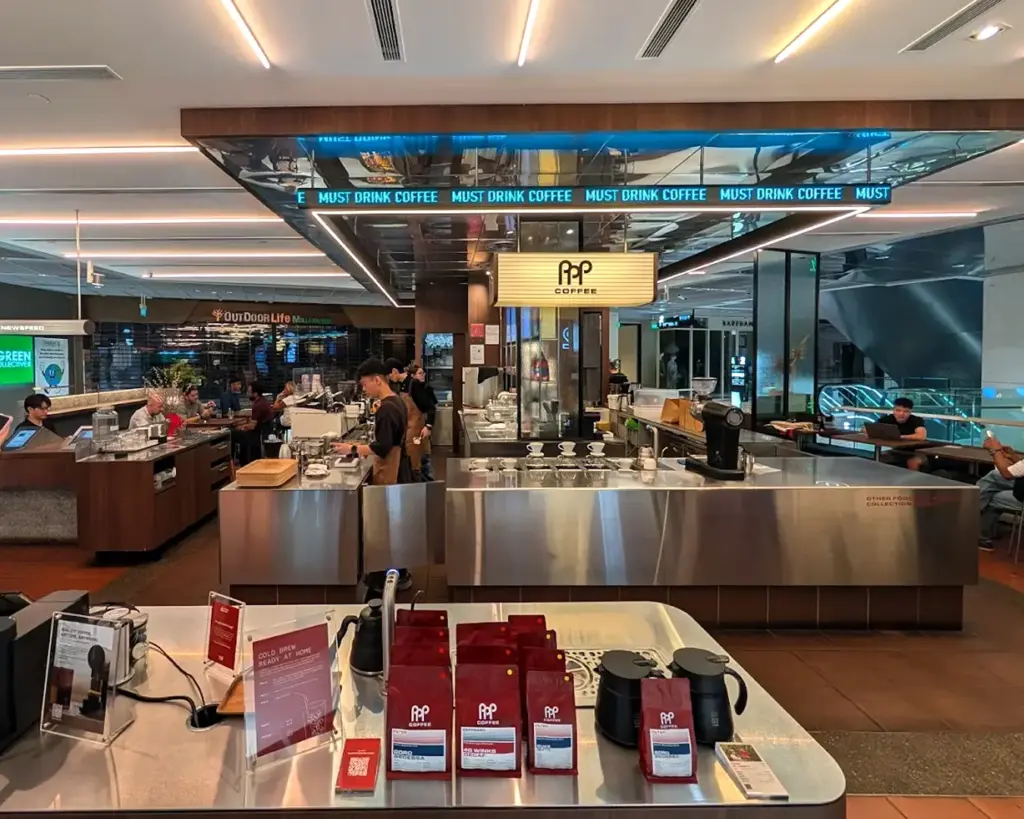 A modern coffee shop interior with a sleek metal counter displaying coffee products. Signs read "Must Drink Coffee." Baristas work behind the counter.