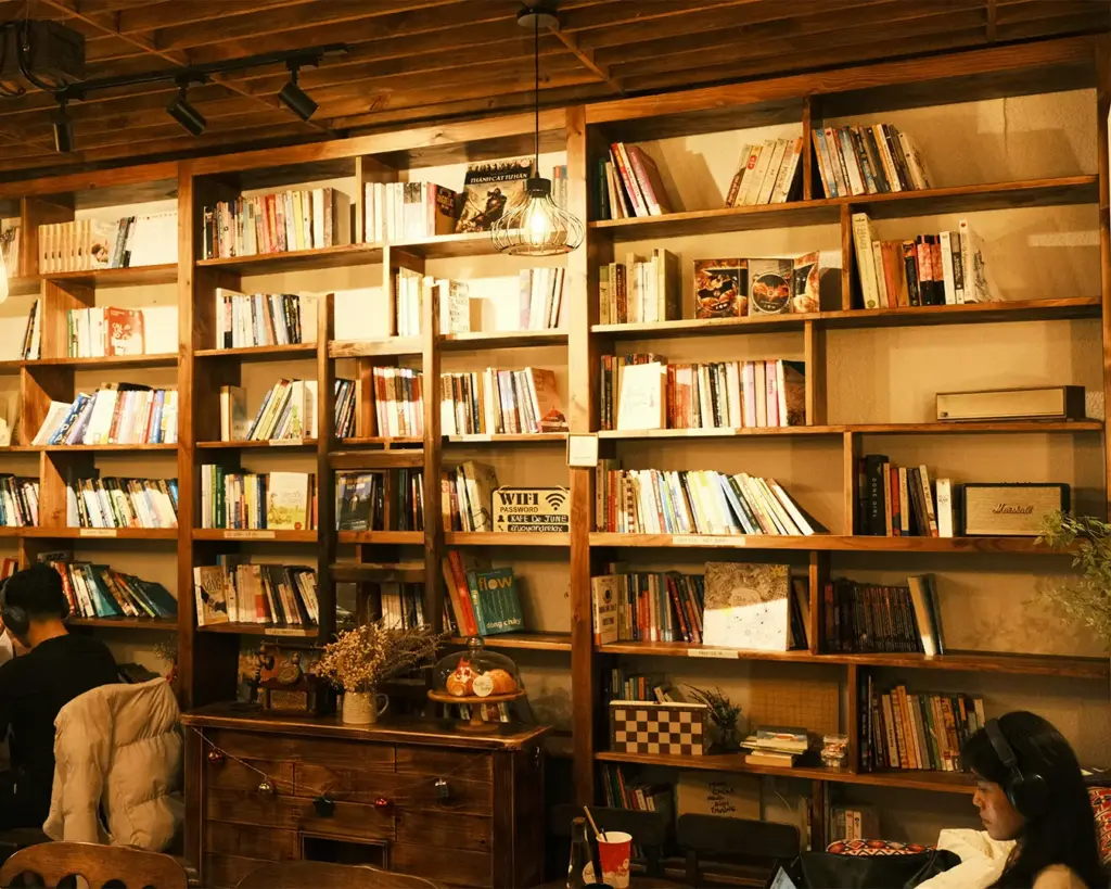 Cozy cafe interior with wooden bookshelves filled with books. Soft lighting creates a warm ambiance. People are sitting, one reading, another with headphones.