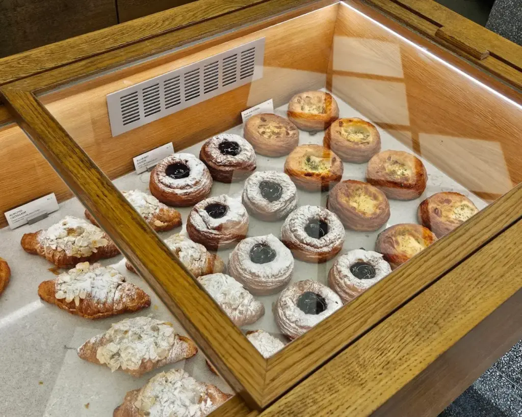 A wooden bakery display case filled with assorted pastries, including almond croissants and powdered pastries with jam centers, creating a tempting, delightful atmosphere.