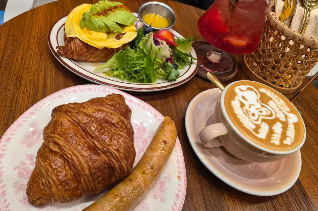 A hearty brunch plate featuring a flaky croissant with scrambled eggs, avocado, a side salad, and a sausage, served alongside specialty coffee and a refreshing drink served at Rise Bakehouse at 111 Somerset.