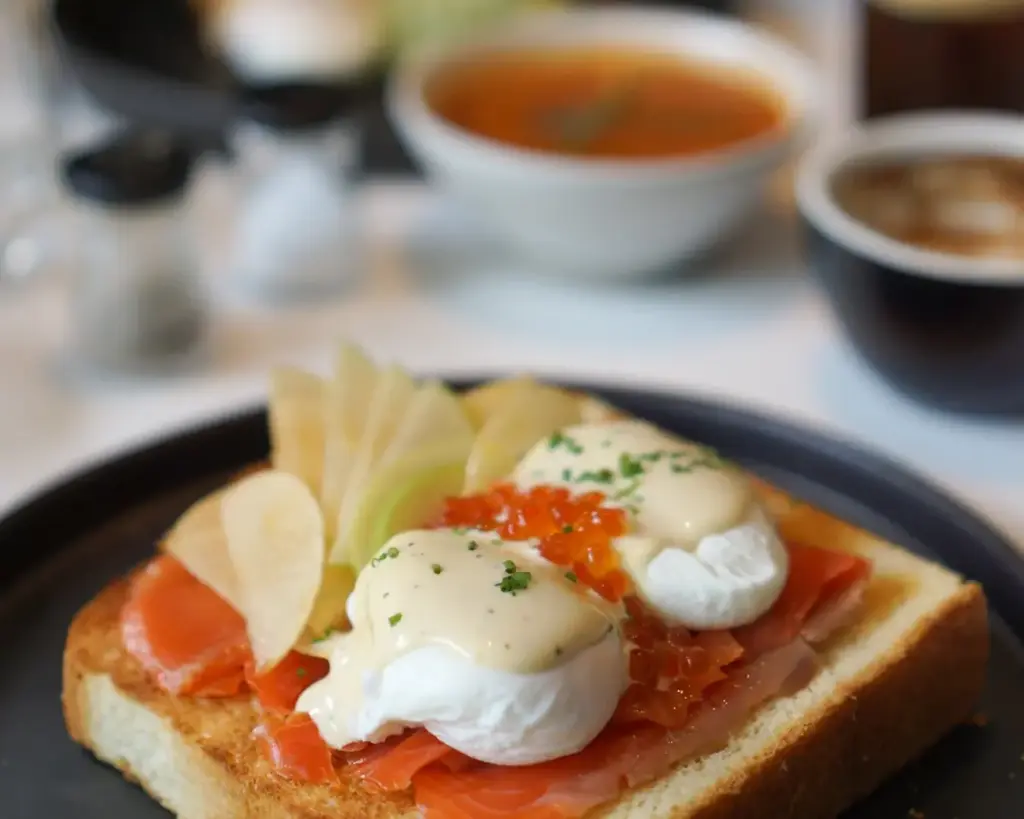 Toast topped with smoked salmon, poached eggs, hollandaise sauce, and herbs, garnished with apple slices. Soup and coffee in the background.