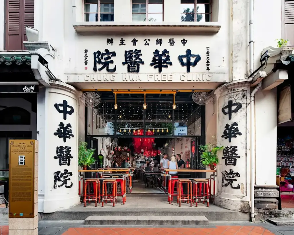 Street-front view of a historic building labeled "Chung Hwa Free Clinic" with bold Chinese characters, tables outside, and a vibrant, cozy atmosphere.