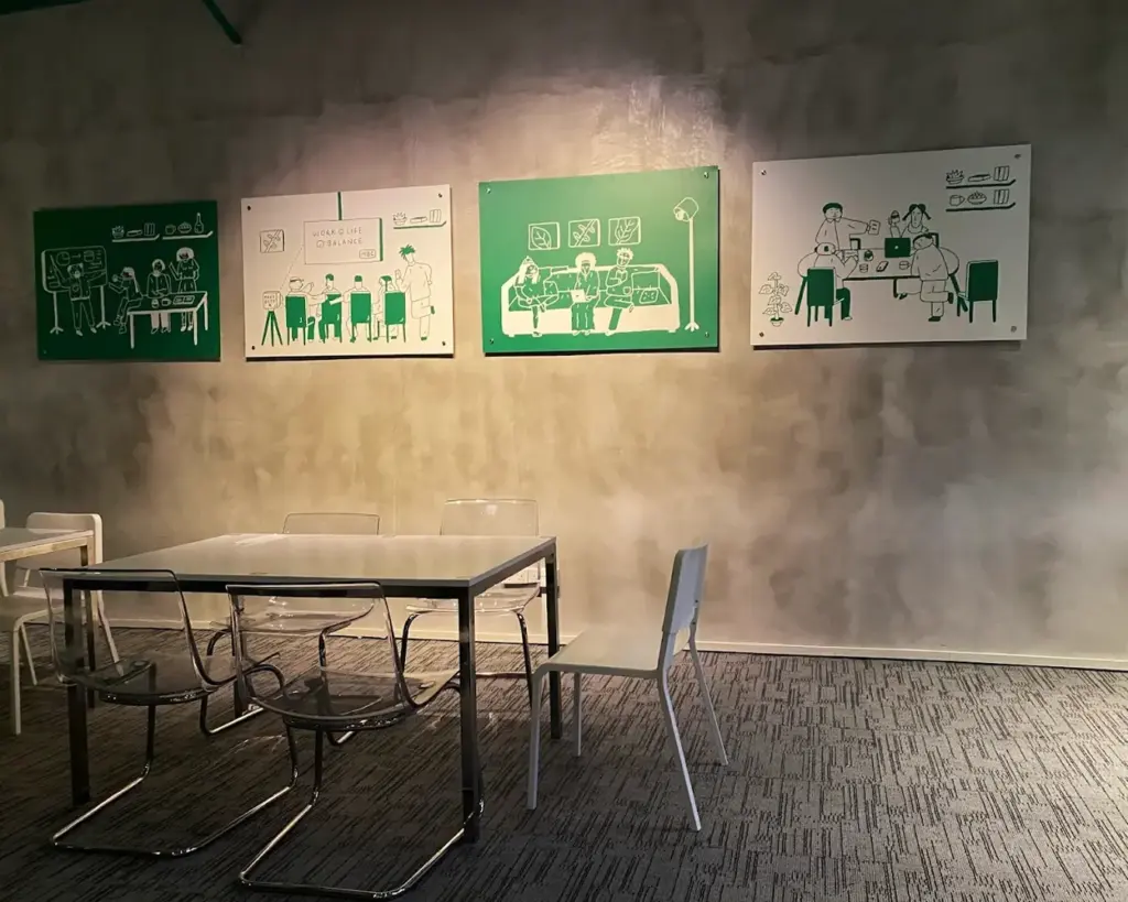 Modern workspace with a white table and transparent chairs. Three green and white illustrations depicting office scenes are mounted on a concrete wall.