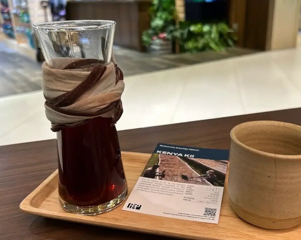 A glass of deep red coffee wrapped in a patterned cloth sits on a wooden tray beside an empty ceramic cup. A card labeled "Kenya Kii" lies nearby.