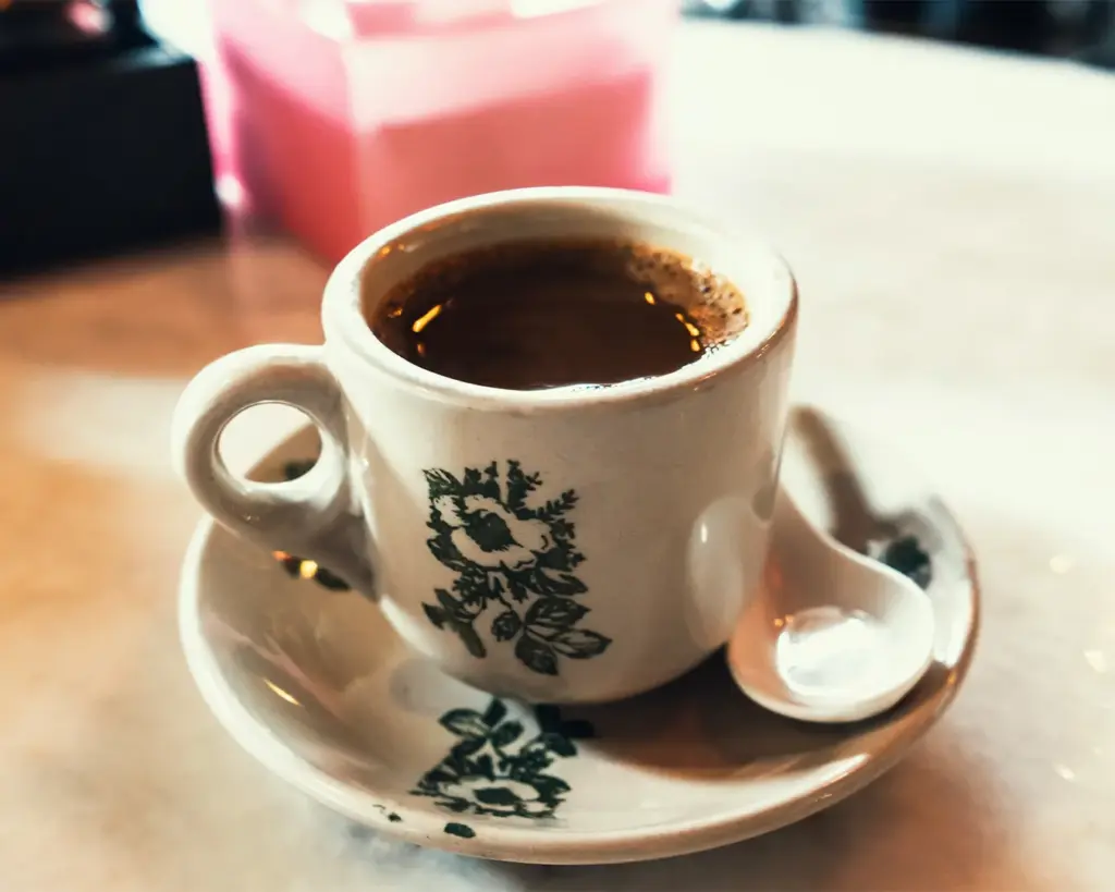 A small white cup of hot coffee with floral design sits on a matching saucer with a spoon. The cozy scene has a warm, inviting atmosphere.