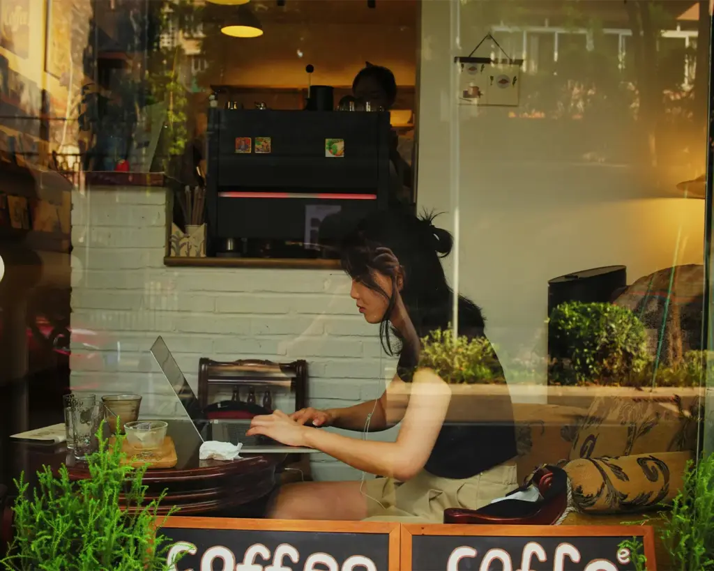 A woman sitting in a cozy cafe working on a laptop by the window. A coffee and glass sit on the table. Warm lighting and greenery create a relaxed ambiance.
