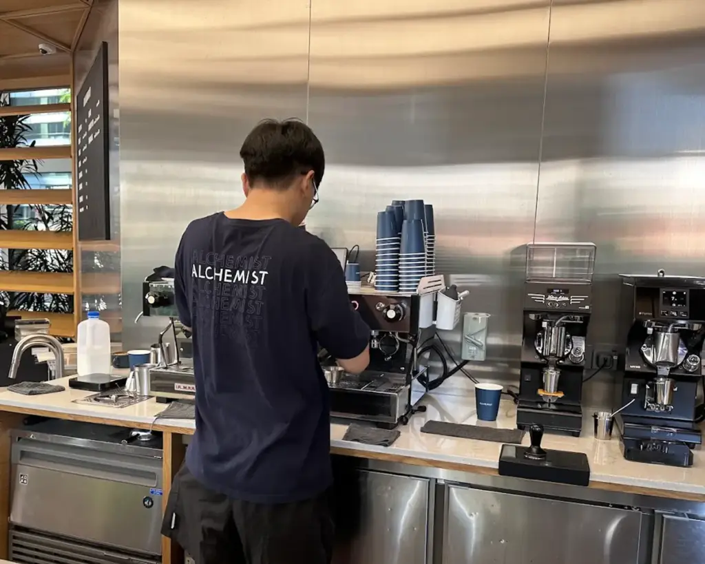 A barista in a navy shirt operates an espresso machine in a modern café setting. Stacked coffee cups and milk carton visible, creating a focused atmosphere.