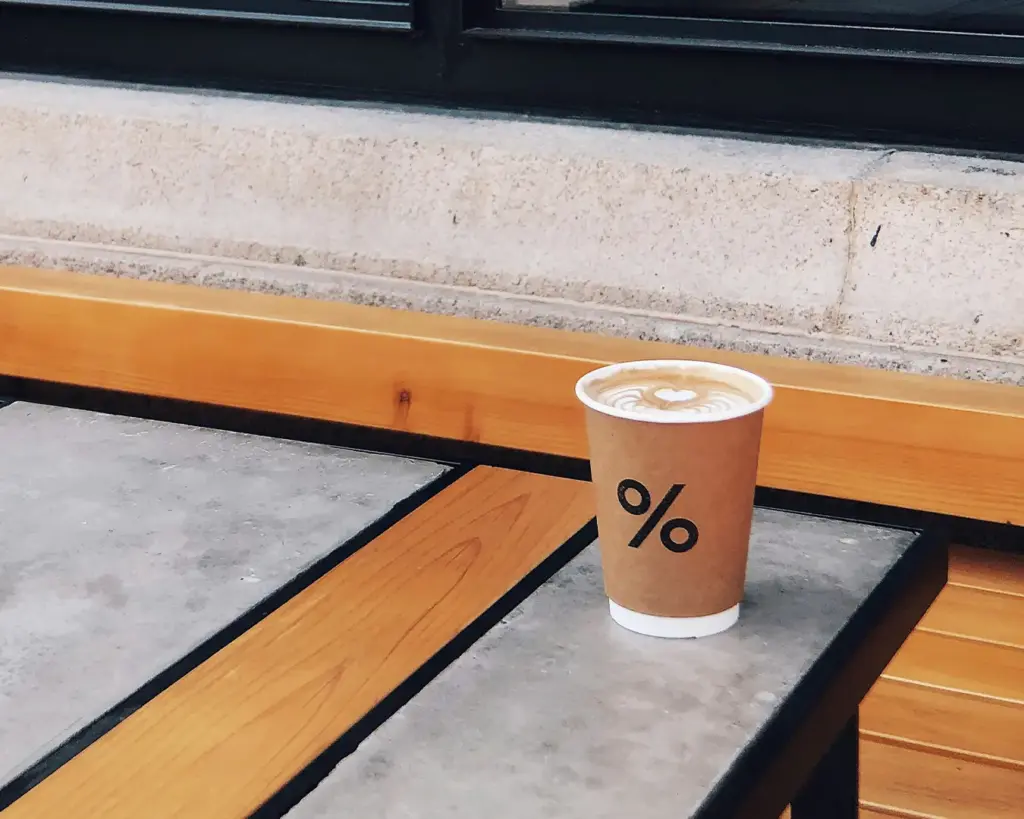 A paper cup with a percent sign logo holds coffee with latte art on top, set on a concrete and wood table, conveying a modern and minimalist vibe.