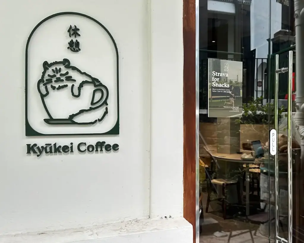 Signage for "Kyūkei Coffee" features a cute cat lounging in a coffee cup. Glass door with promo poster leads into a cozy café interior.