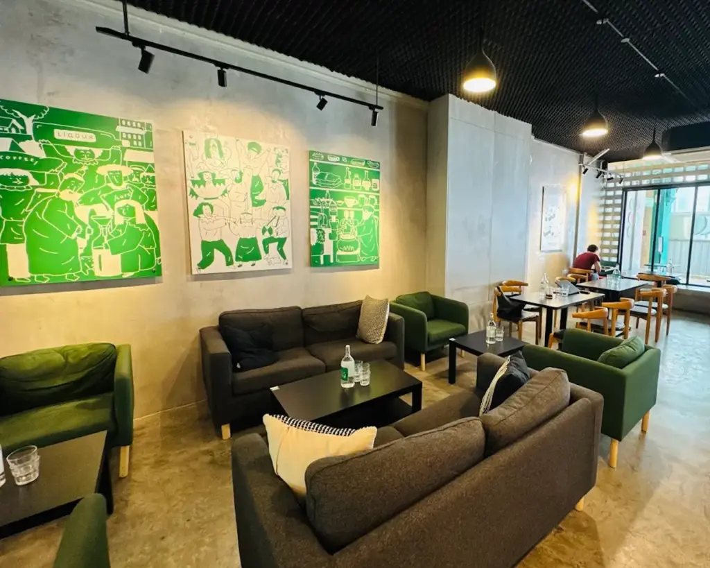 Cozy cafe interior with green and gray couches, wooden chairs, and modern art on concrete walls. Soft lighting and relaxing ambiance.