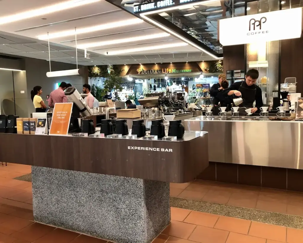 Modern coffee shop with an "Experience Bar," featuring kettles and a barista pouring a drink. Customers chat nearby. Bright, inviting atmosphere.