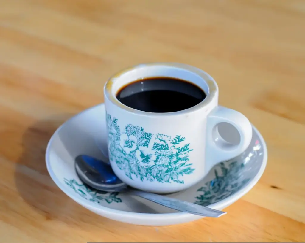 A white ceramic cup filled with black coffee sits on a matching saucer with green floral patterns. A silver spoon rests beside it on a light wooden table.
