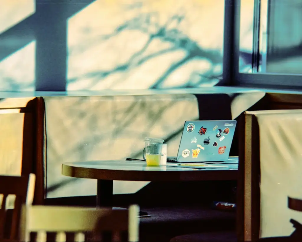 A sunlit café booth with a table; a sticker-covered laptop and a half-full lemonade glass are present. Tree shadows create a serene atmosphere.