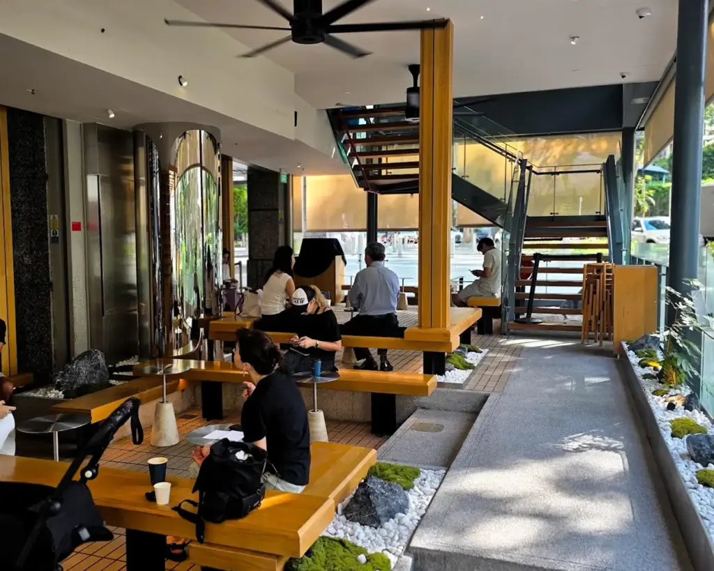 Modern café interior with wooden benches and tables, patrons relaxing. Sunlight streams through large windows, illuminating the serene space. Cozy ambiance.