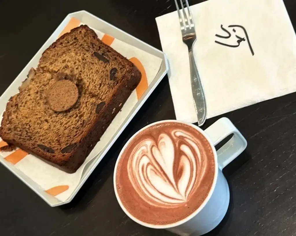 A slice of banana bread with a round biscuit on top sits on a tray. Next to it, a cup of hot chocolate features latte art, adjacent to a fork and a napkin.