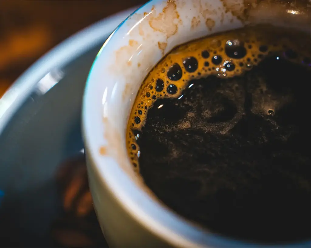 Close-up of a steaming cup of black coffee with rich crema on top. The cup's rim shows coffee stains, conveying a warm and inviting tone.