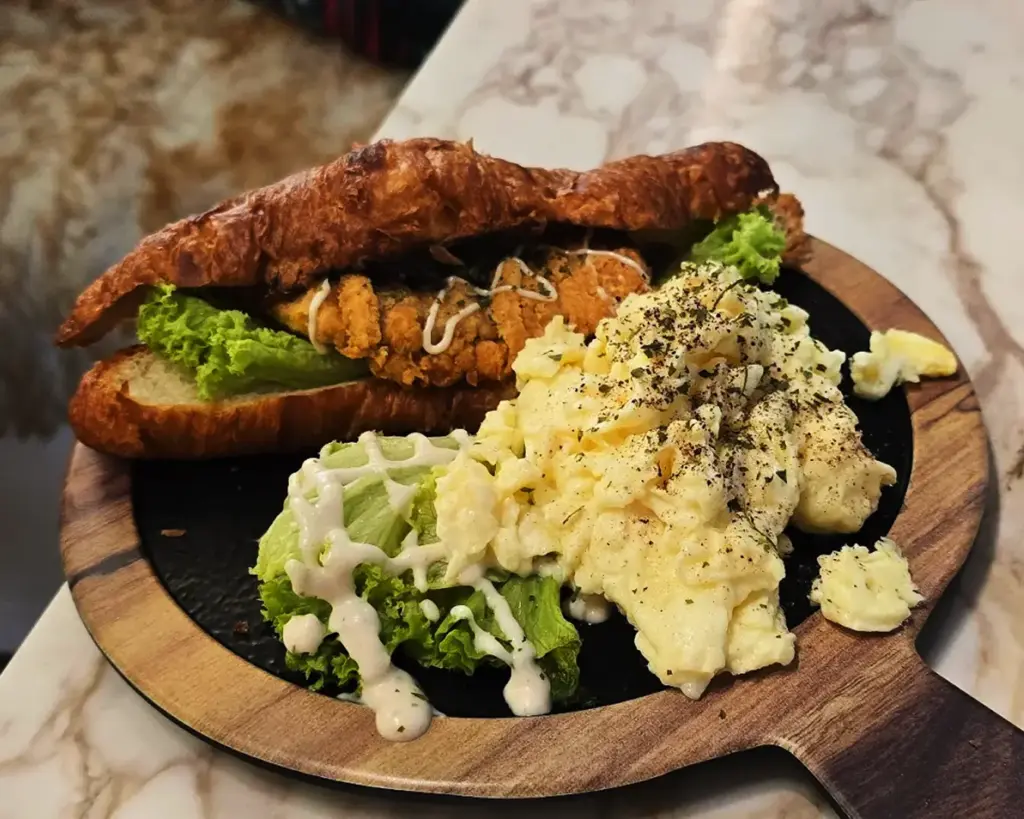 A croissant sandwich with fried chicken and lettuce, drizzled with sauce, accompanied by seasoned scrambled eggs and lettuce, on a wooden serving board.