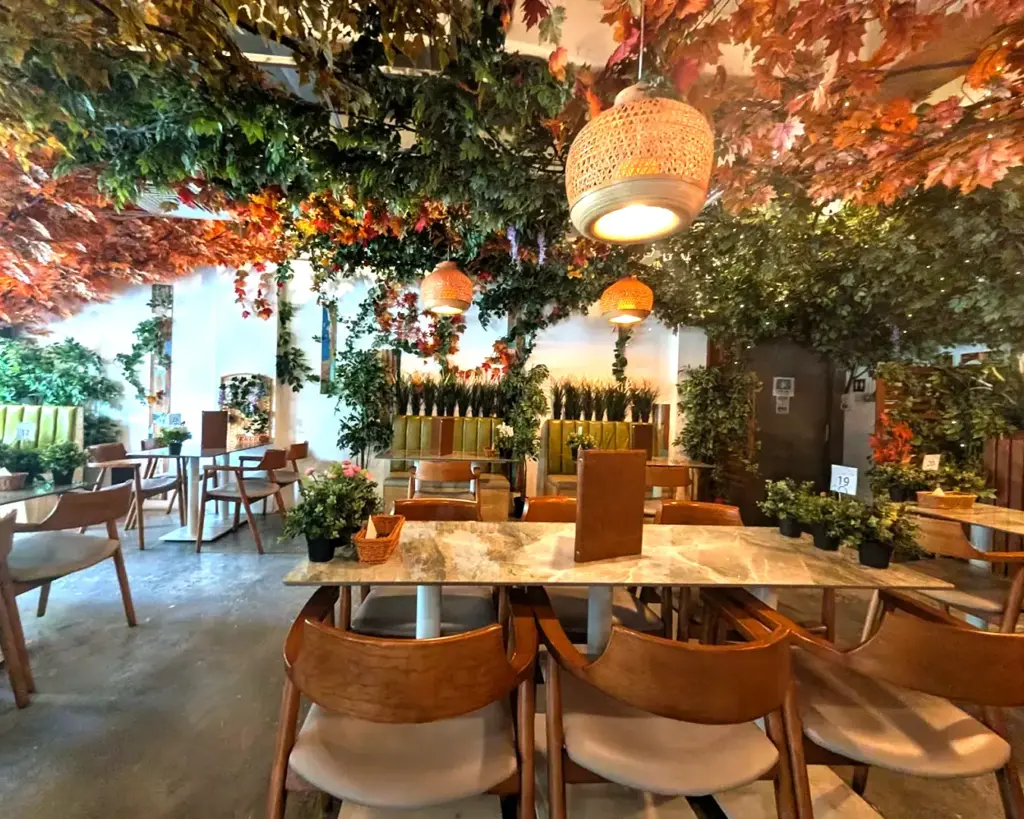 Cozy cafe interior with wooden chairs and tables. Lush green and autumn leaves hang from the ceiling, creating a warm, inviting atmosphere.