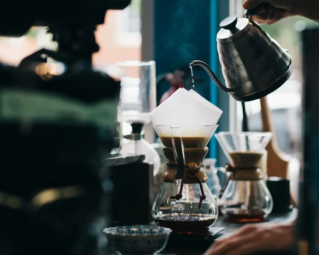 A person pours hot water from a gooseneck kettle into a Chemex, brewing coffee with a serene, focused atmosphere. Steam rises from the filter.