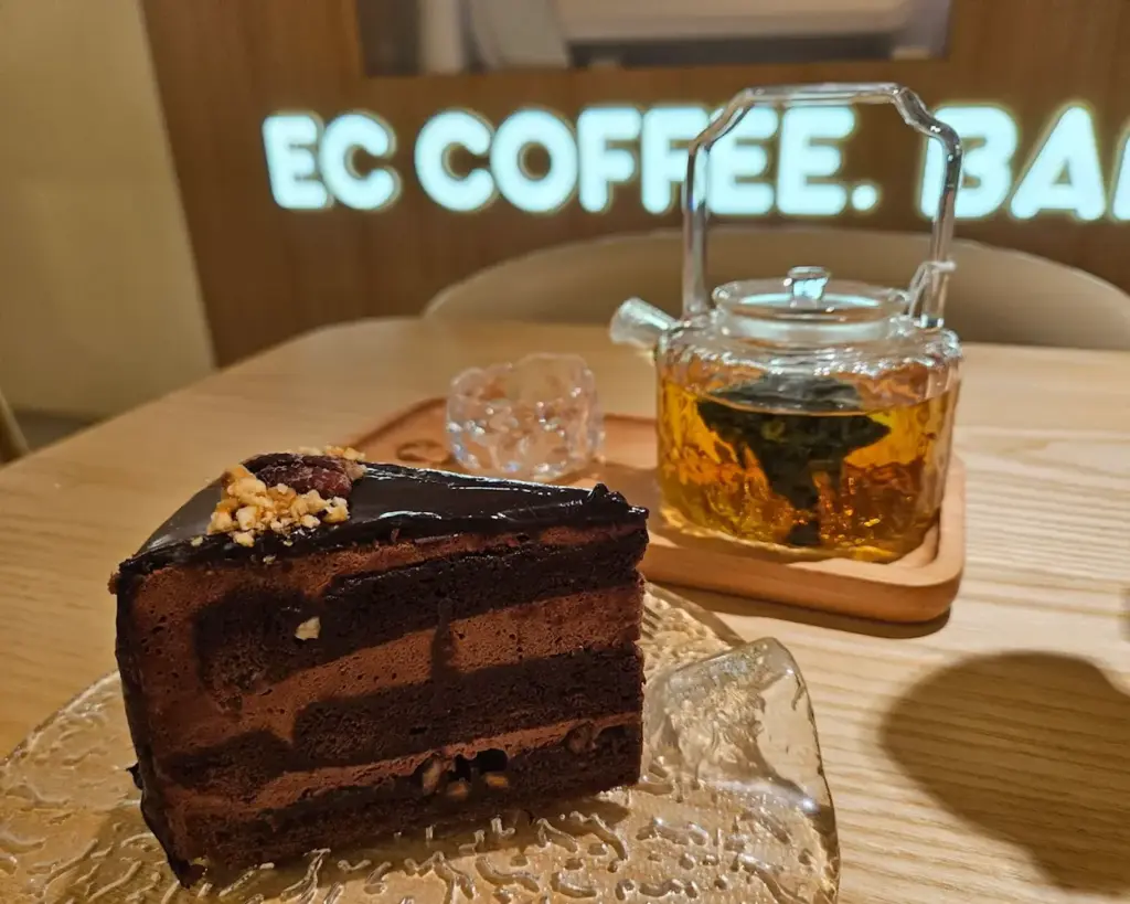 A slice of rich chocolate cake with layers and nuts on a clear plate, paired with a glass teapot of herbal tea on a wooden table. Warm, cozy ambiance.