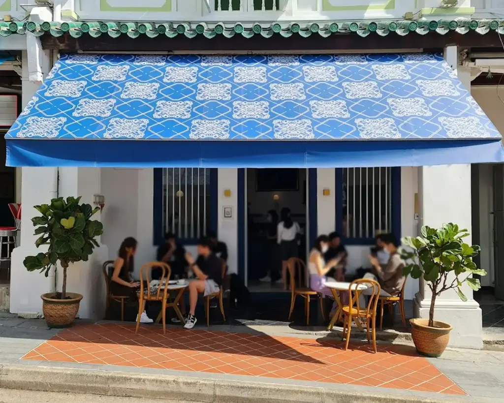 Front view of a cafe with a blue patterned awning, people sitting at wooden chairs and tables outside, surrounded by potted plants on a sunny day.