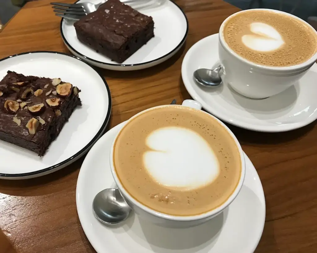 Cappuccinos with heart-shaped latte art on a wooden table, surrounded by two chocolate brownies, one topped with hazelnuts. Cozy cafe atmosphere.