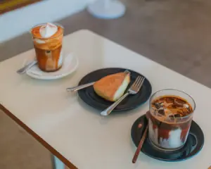 Two glasses of iced coffee with milk and caramel sit on a white table, beside a slice of cake on a plate with a fork. The setting is casual and inviting.