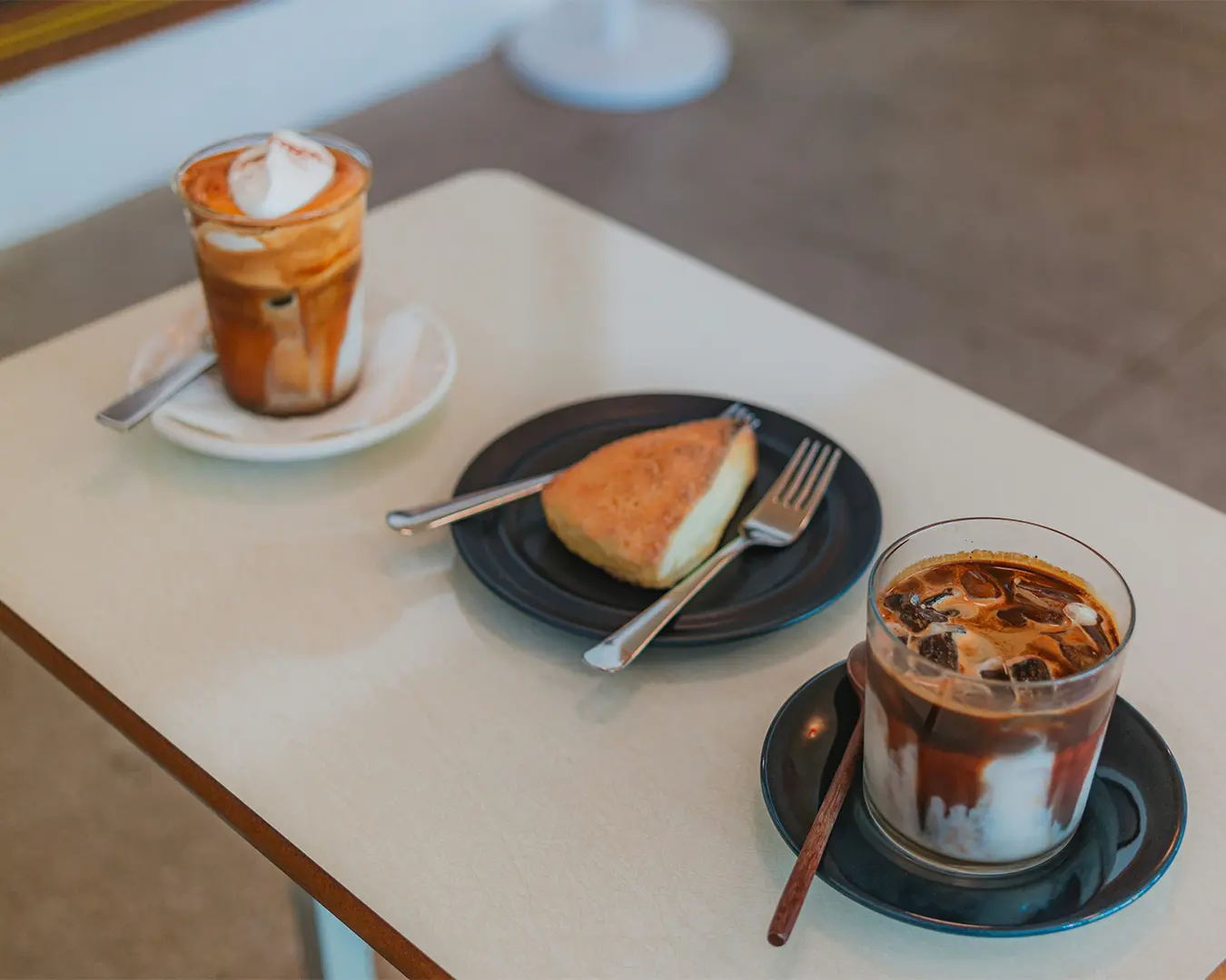Two glasses of iced coffee with milk and caramel sit on a white table, beside a slice of cake on a plate with a fork. The setting is casual and inviting.