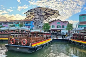 Traditional Singapore river cruise boats docked at Clarke Quay, featuring colorful colonial-style shops and a large modern canopy structure under a blue sky.