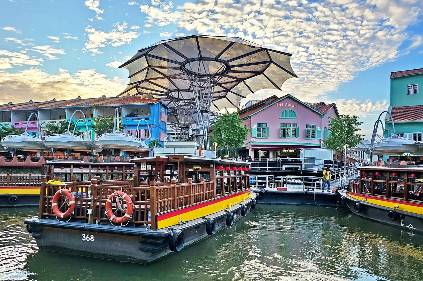 Traditional Singapore river cruise boats docked at Clarke Quay, featuring colorful colonial-style shops and a large modern canopy structure under a blue sky.