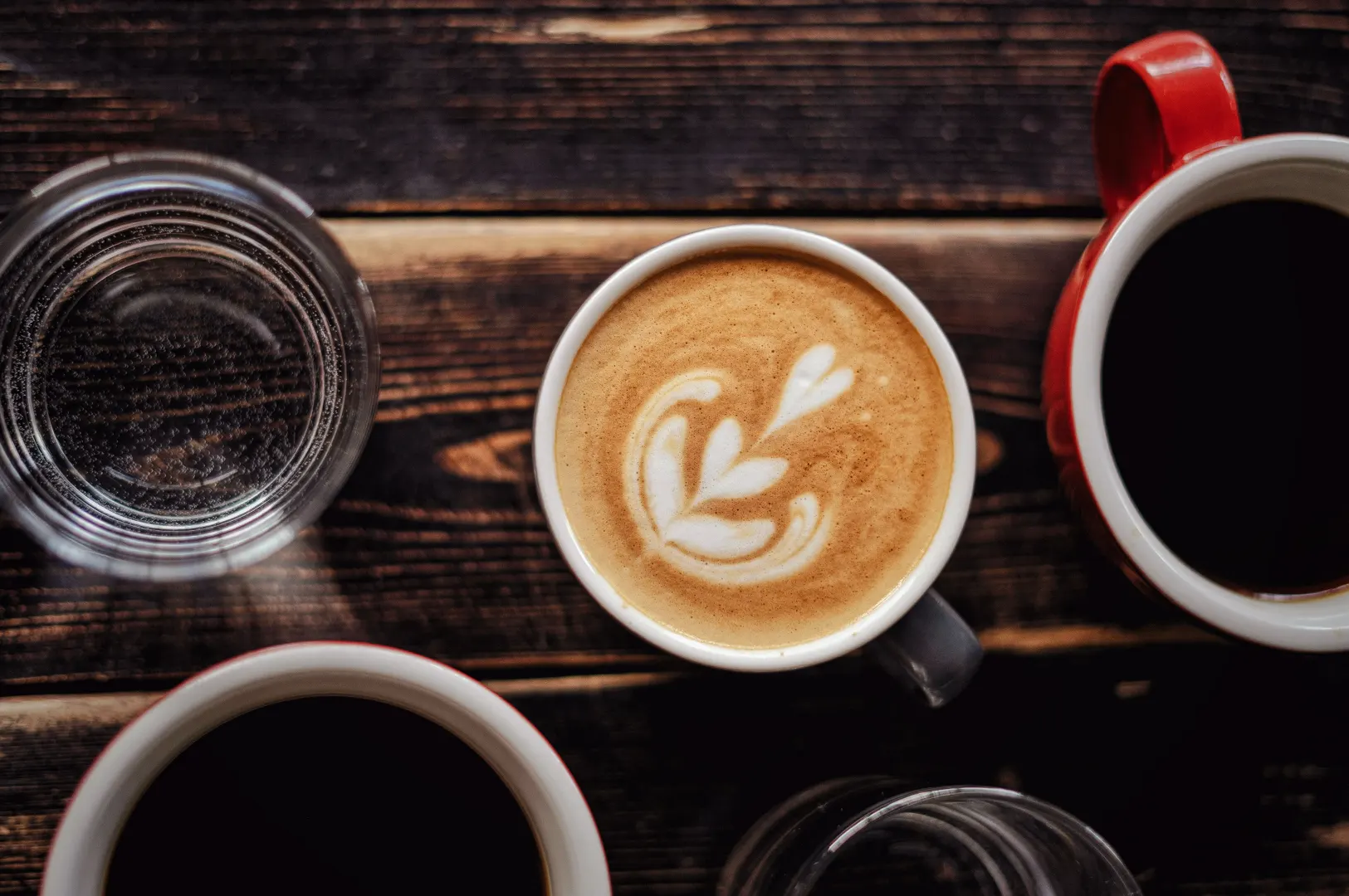 Top-down view of a cup of latte art coffee alongside black coffee and glasses of water on a rustic wooden table, representing Singaporean coffee culture.