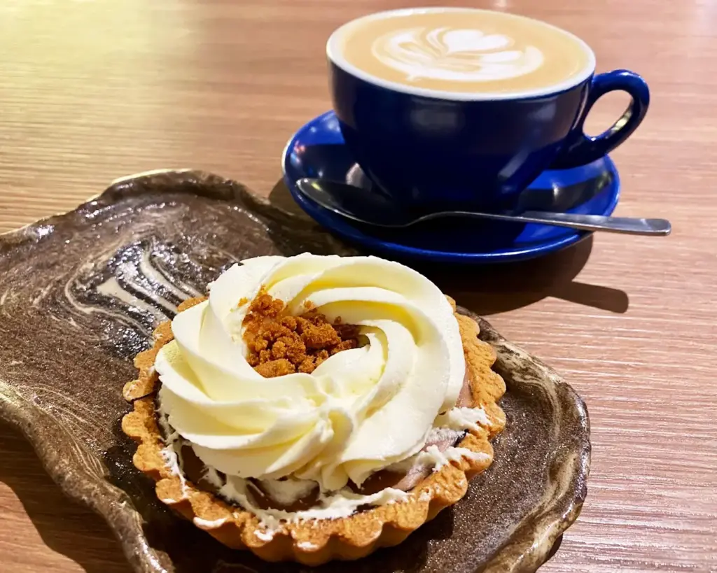 A beautifully plated tart topped with whipped cream and crumbs sits beside a blue cup of latte with elegant foam art, creating a cozy café vibe.