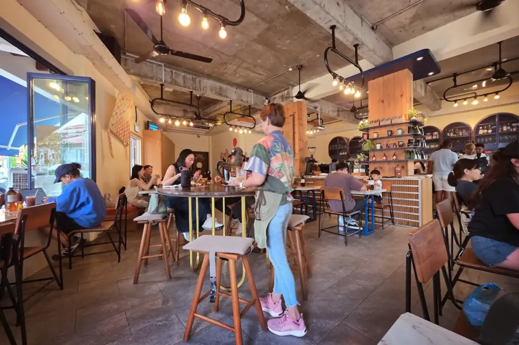 Patrons dining at the industrial-chic interior of Common Man Coffee Roasters at Joo Chiat Road, showcasing the café's bustling atmosphere, the comfortable seating mix for different groups, and the coffee bar.