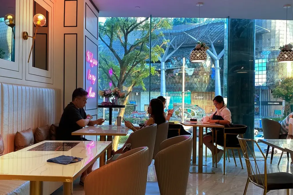 Patrons seated in a modern, well-lit café with floor-to-ceiling windows overlooking a city center, showcasing a prime example of a professional and comfortable work-friendly café culture in Singapore.