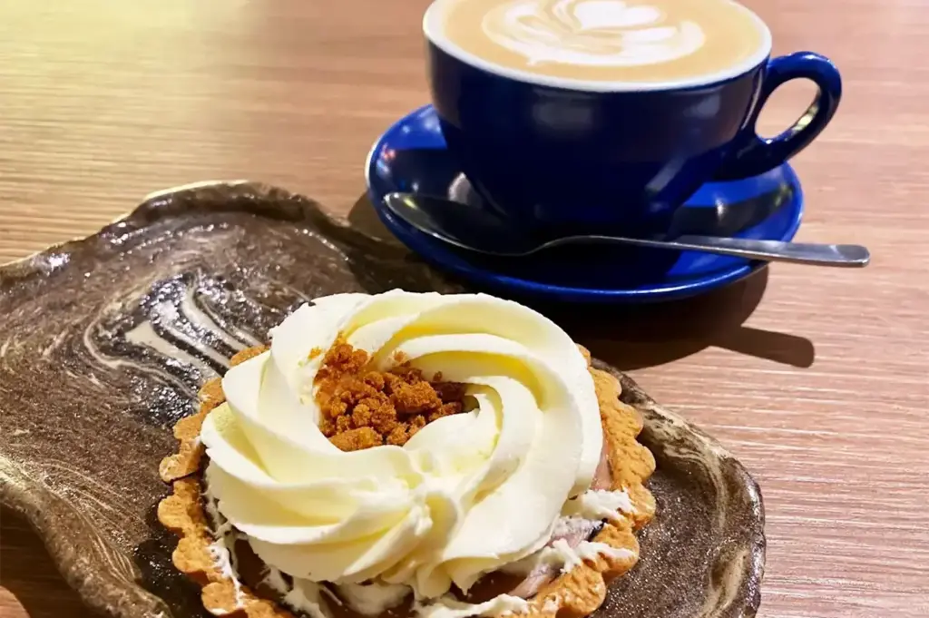 A beautifully plated tart topped with whipped cream and crumbs sits beside a blue cup of latte with elegant foam art, creating a cozy café vibe.