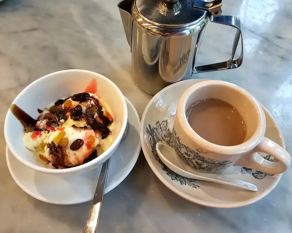 A bowl of ice cream with syrup and toppings sits next to a cup of coffee with a saucer, spoon, and metal pot on a marble surface.