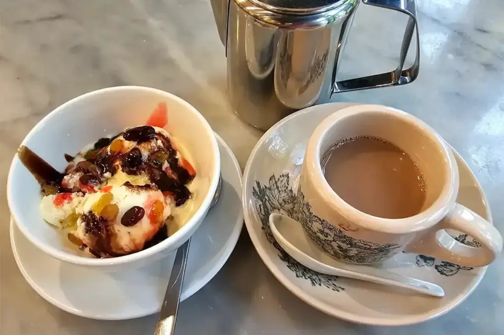 A bowl of ice cream with syrup and toppings sits next to a cup of coffee with a saucer, spoon, and metal pot on a marble surface.
