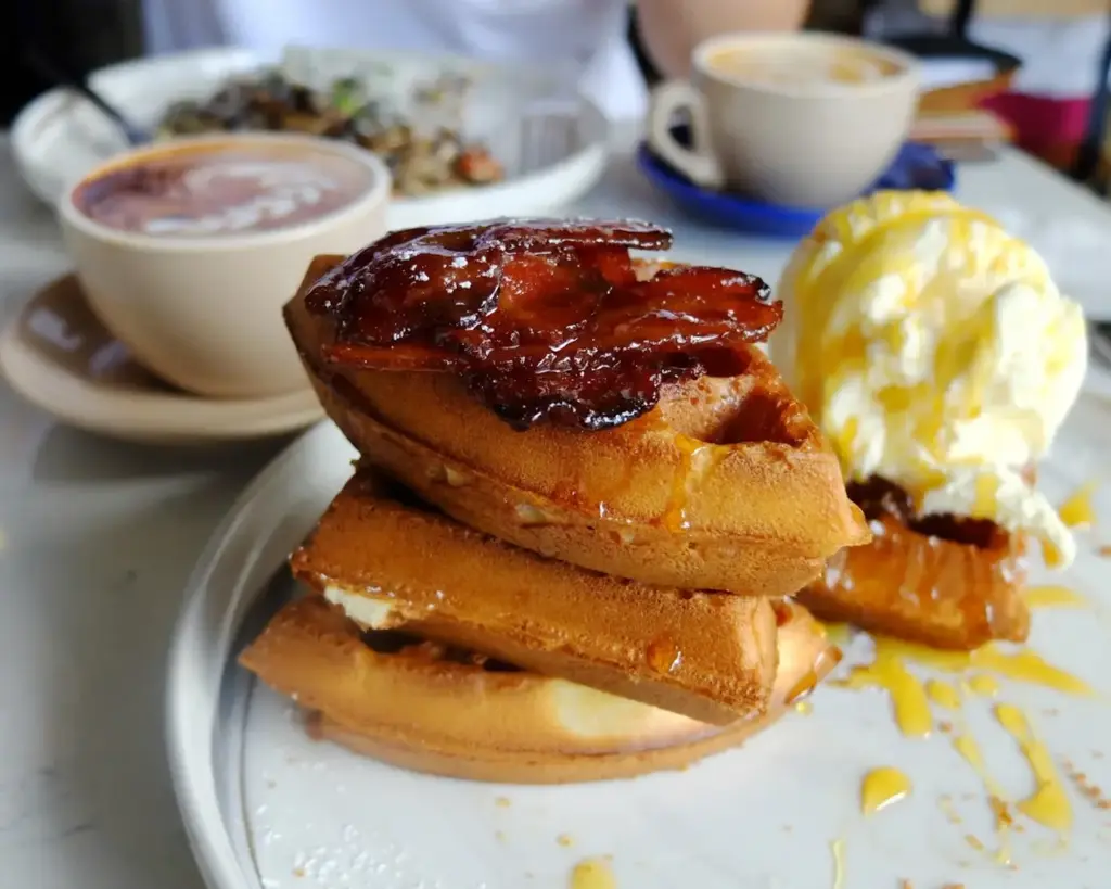 Stack of golden waffles topped with candied bacon and served with vanilla ice cream, syrup, and coffee in the background.