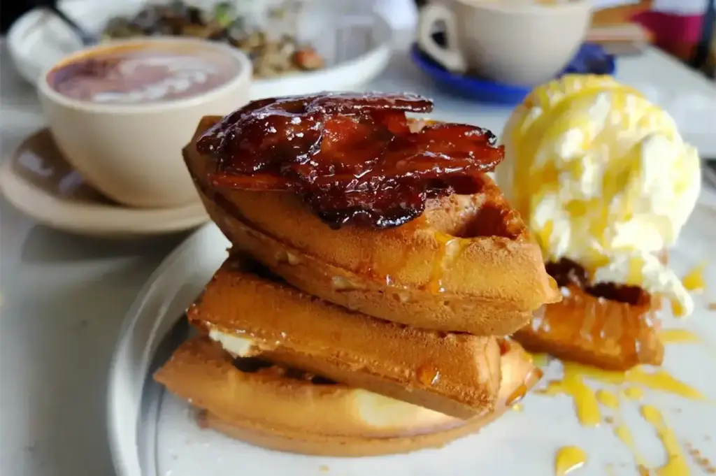 Stack of golden waffles topped with candied bacon and served with vanilla ice cream, syrup, and coffee in the background.