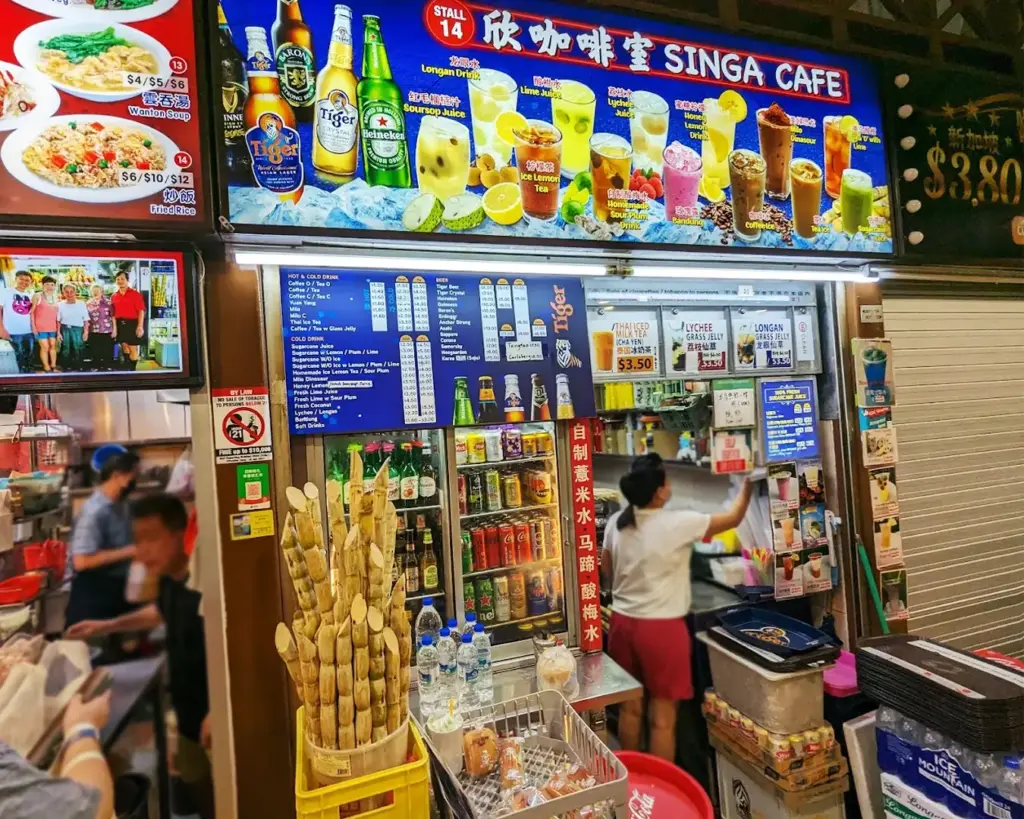 Brightly lit Singapore hawker café stall displaying colorful drink menus, bottled beverages, sugarcane stalks, and iced local drinks.