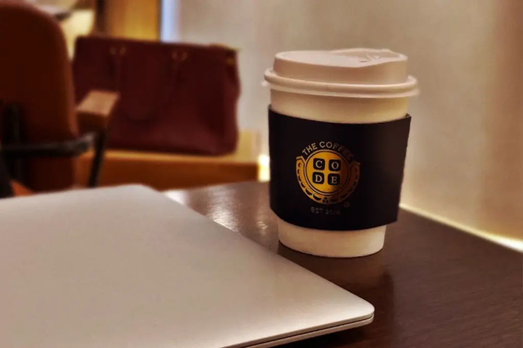 A professional workspace setup inside The Coffee Code Singapore, featuring a laptop on a wooden table next to a takeaway cup branded with The Coffee Code logo.