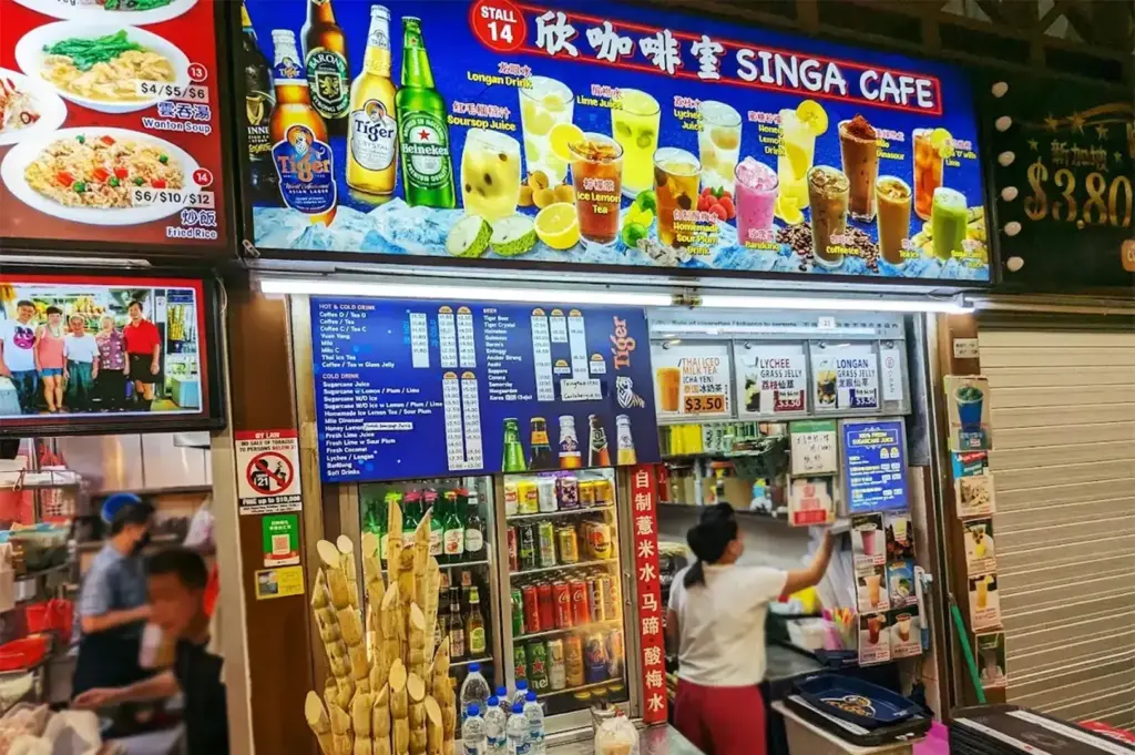 Brightly lit Singapore hawker café stall displaying colorful drink menus, bottled beverages, sugarcane stalks, and iced local drinks.