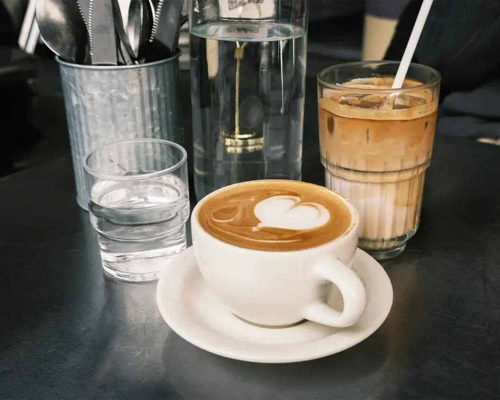 Close-up of a creamy cappuccino with heart-shaped latte art beside an iced latte, glass of water, and metal utensil holder on a dark café countertop. The warm tones and coffee presentation highlight a stylish specialty coffee shop environment.