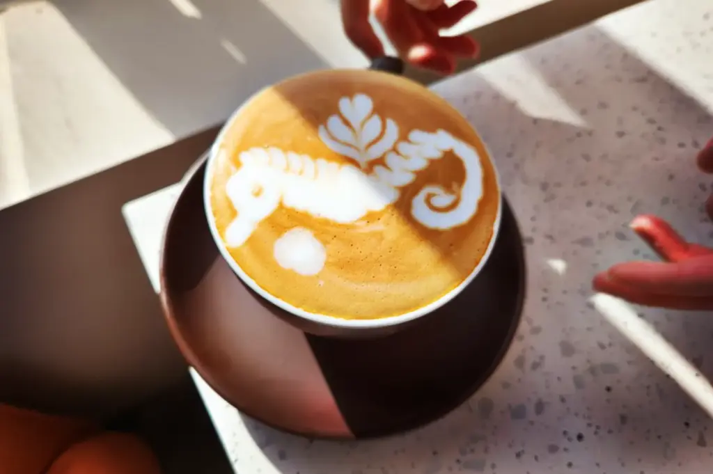 A close-up of a cup of coffee featuring intricate seahorse-shaped latte art, served on a brown plate on a bright, modern tabletop.