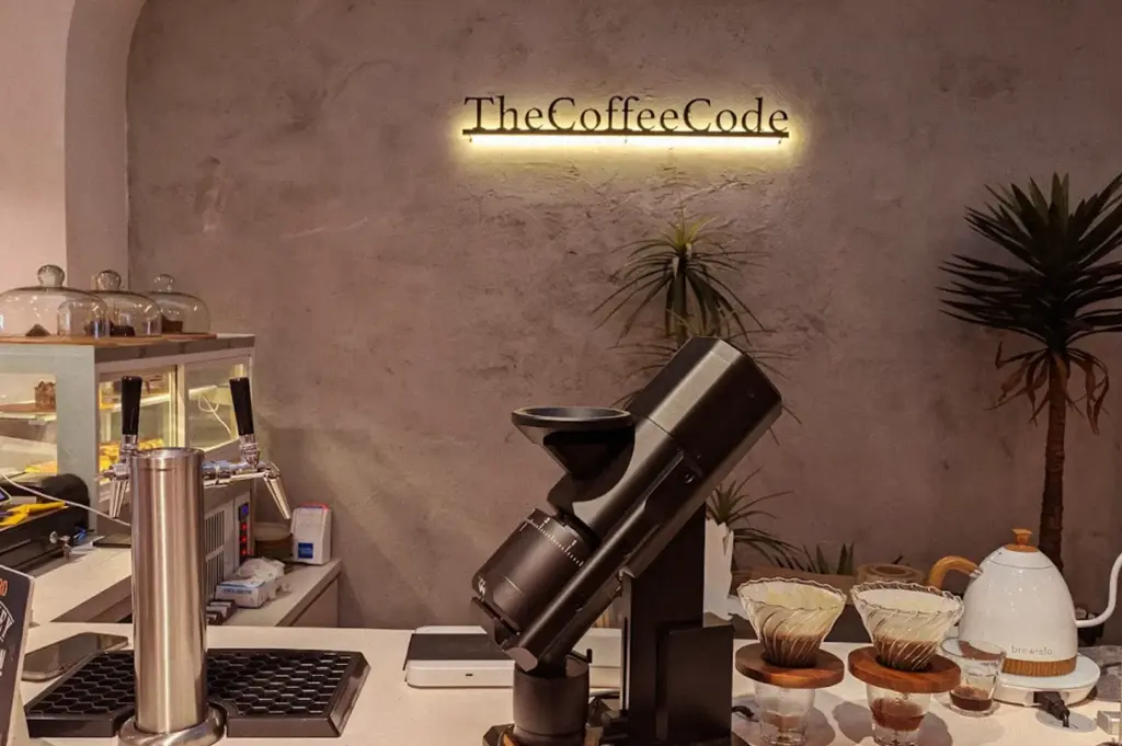 A close-up of the coffee brewing station at The Coffee Code Singapore, featuring a professional grinder, a white gooseneck kettle, and the cafe's illuminated sign on a textured grey wall.